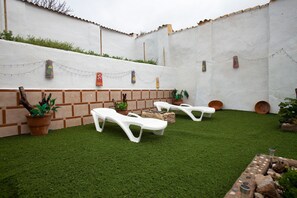 Une cour avec une pelouse artificielle, des fauteuils blancs, des plantes en pot et des éléments décoratifs suspendus au mur.