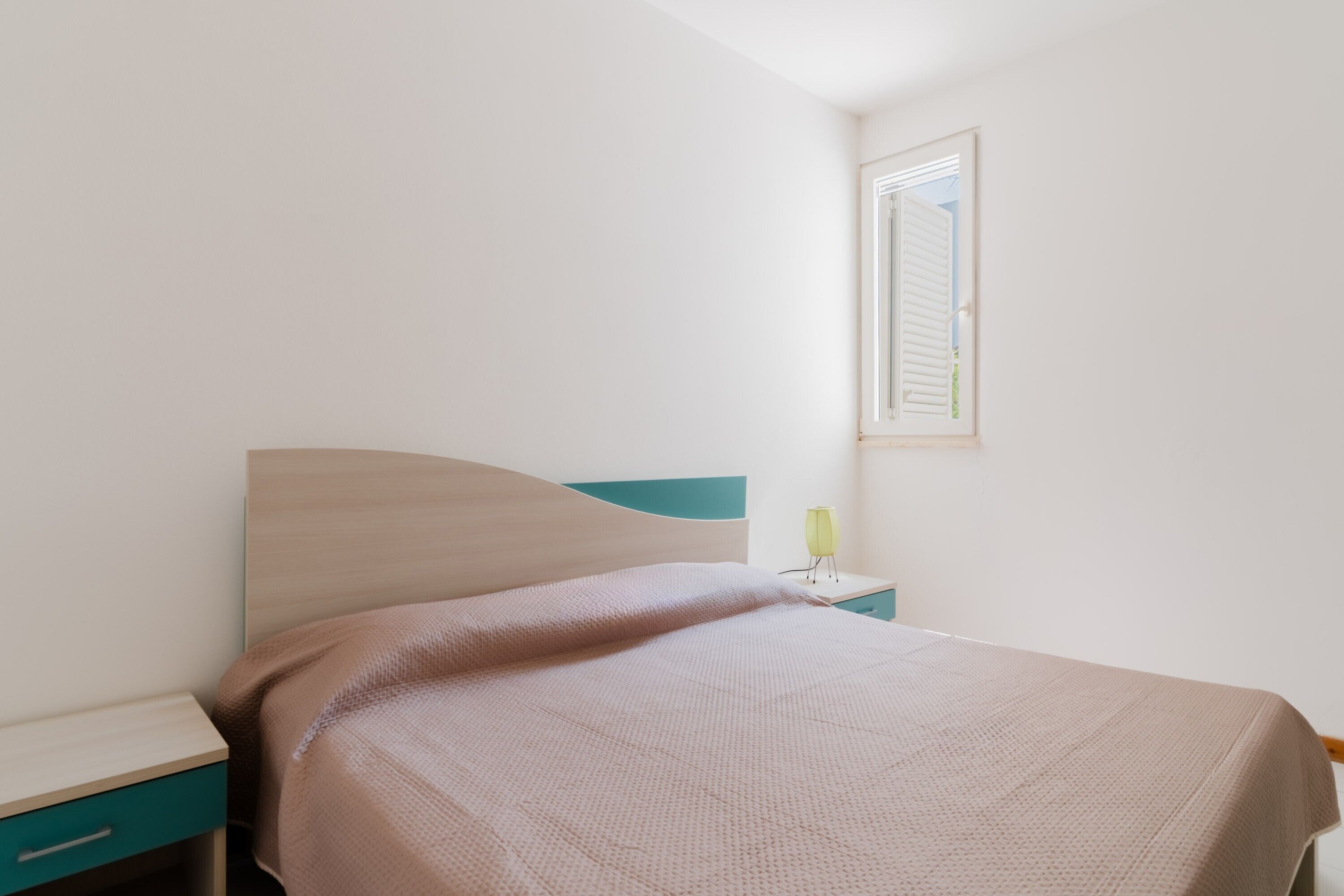 Un dormitorio con una cama, una mesita de noche, un cajón y una ventana con persianas.