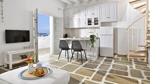 A modern kitchen with white cabinets, a built-in island, two black chairs, a white refrigerator, and a TV mounted on the wall. - Apartment 'Casa D'irene Mykonian Residence' with Sea View, Wi-Fi and Air Conditioning (Mýkonos City)