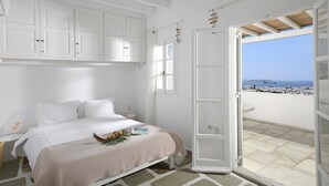A bedroom with a bed, white cabinets, a nightstand, a window, and an open door leading to a balcony with a view of the sea. - Apartment 'Casa D'irene Mykonian Residence' with Sea View, Wi-Fi and Air Conditioning (Mýkonos City)
