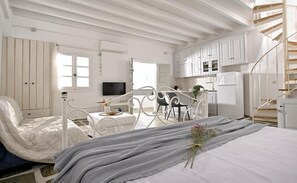 Un dormitorio con cama, una silla, una mesita, televisión y una zona de cocina con armarios y un refrigerador.