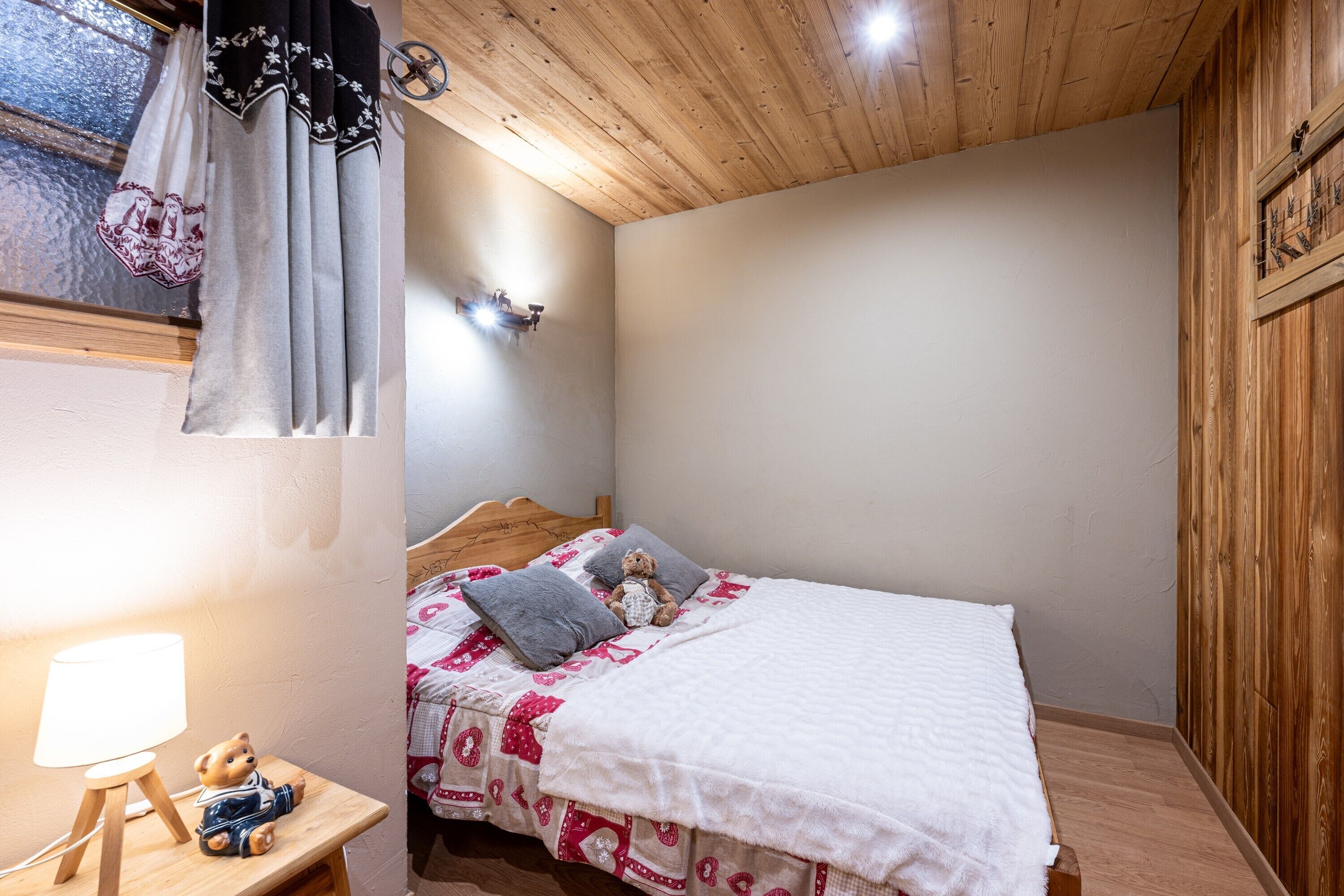 A bedroom with a wooden ceiling, a bed with a white bedspread and pillows, a bedside table with a lamp, and a teddy bear.