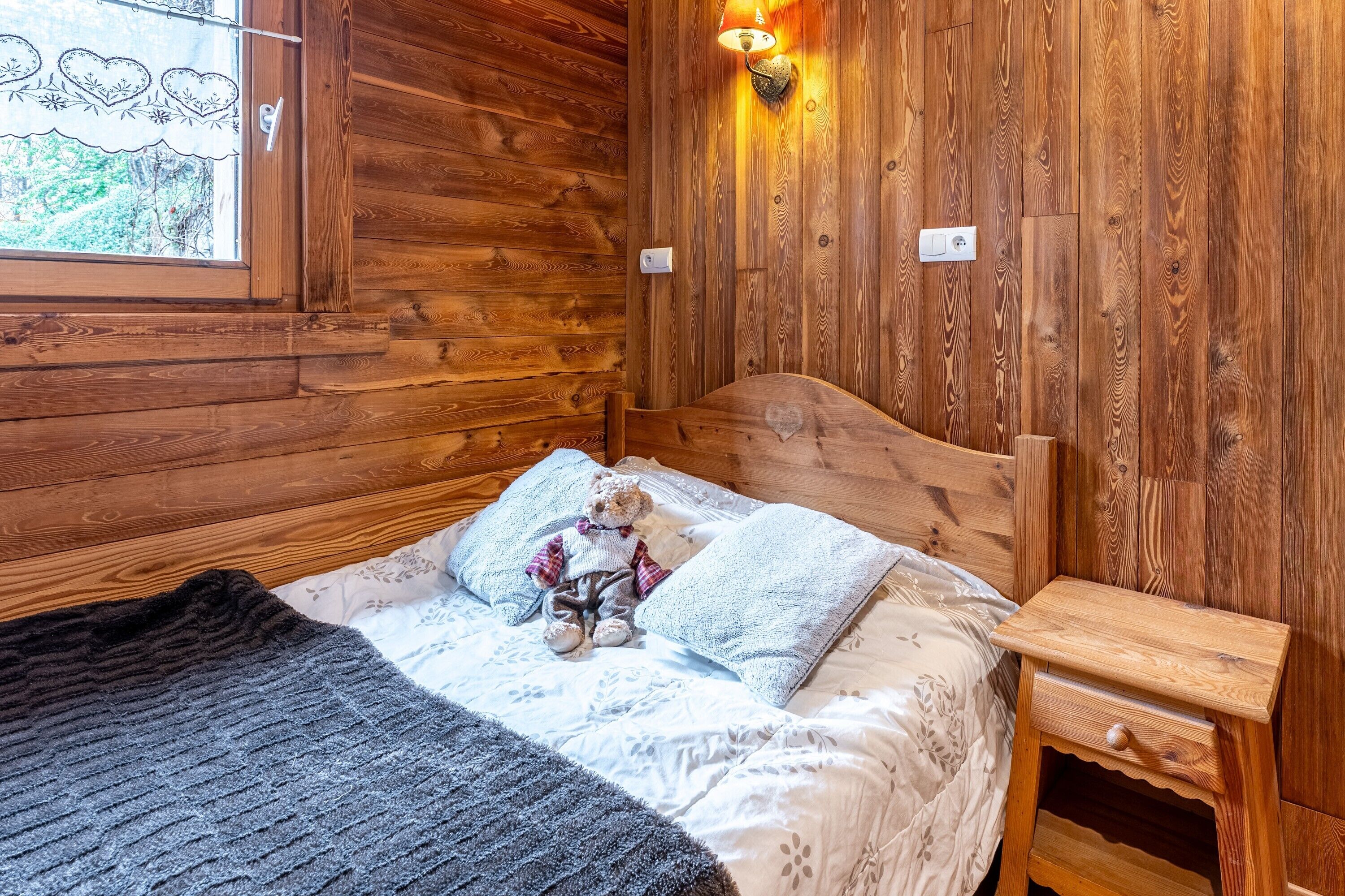 Une chambre dans un chalet en bois, avec un lit, une table de chevet, un ours en peluche et une fenêtre donnant sur la forêt.