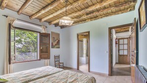 Un dormitorio con techo de madera, un ventanal grande, una cama con colcha estampada, una silla y una puerta que da a otra habitación.