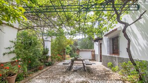 Un patio adoquinado con una mesa y sillas, rodeado de plantas en macetas y una pérgola.