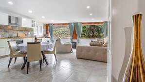 Eine moderne Küche mit weißen Schränken, ein Essbereich mit einem runden Tisch und Stühlen, eine Couch und Blick ins Grüne durch große Fenster.