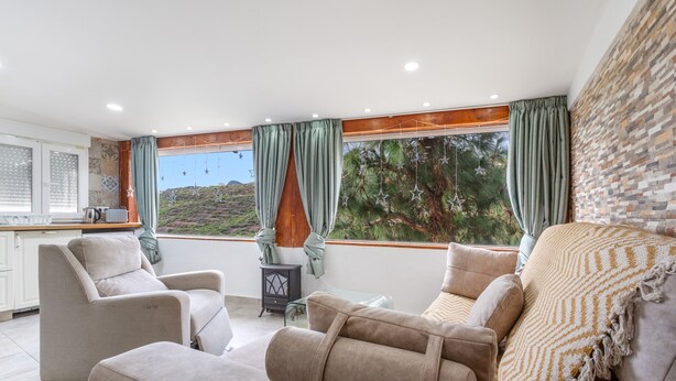 Ein modernes Wohnzimmer mit einer Couch, einem Sessel, einem Hocker, einem kleinen Beistelltisch und Blick auf eine Berglandschaft durch große Fenster.