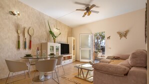 Un salon moderne avec une table à manger en verre, deux chaises, un canapé, un ventilateur de plafond et une porte-fenêtre coulissante donnant sur un balcon.