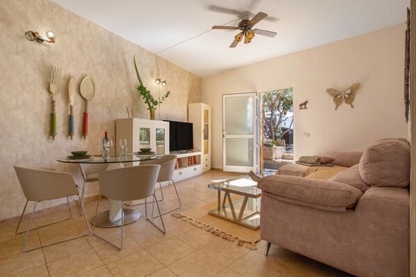 A modern living room with a glass dining table, two chairs, a sofa, a ceiling fan, and a sliding glass door leading to a balcony.