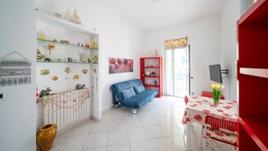 Una habitación con un radiador blanco, un sofá azul, una estantería roja, una mesa con mantel rojo y un jarrón con flores.
