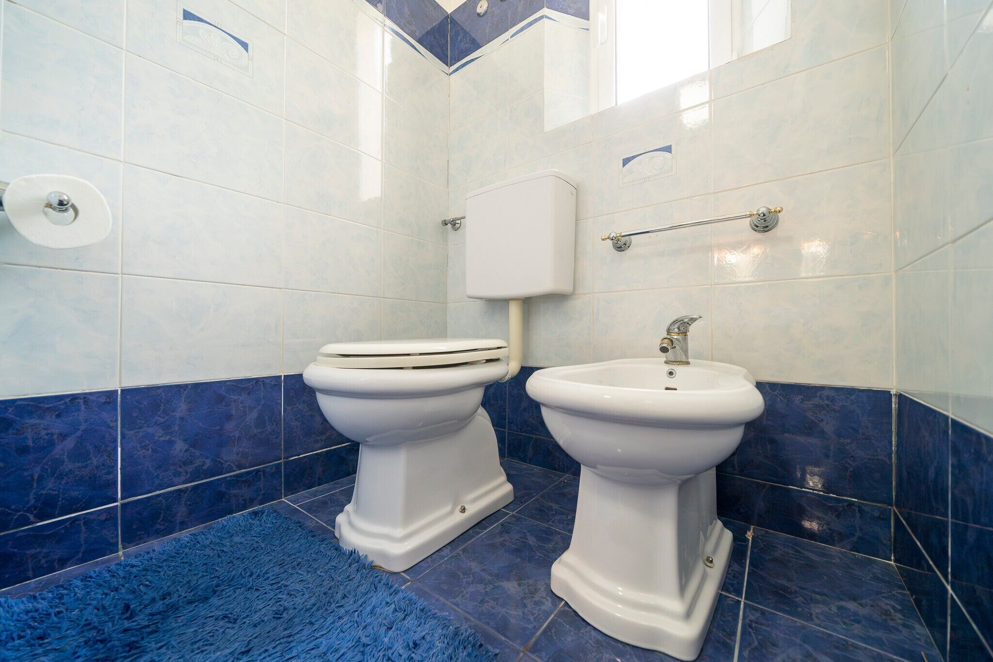 A bathroom with two toilets, a bidet, and a blue rug.