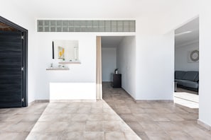 Ein modernes Interieur mit Fliesenboden, einer schwarzen Tür, einem Flur, der in einen Wohnbereich führt, und einem an der Wand befestigten Regal mit Dekorationselementen.