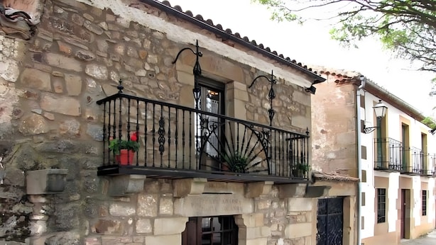 Un edificio de piedra con una puerta de madera, un balcón con barandilla de hierro forjado y una pequeña maceta.