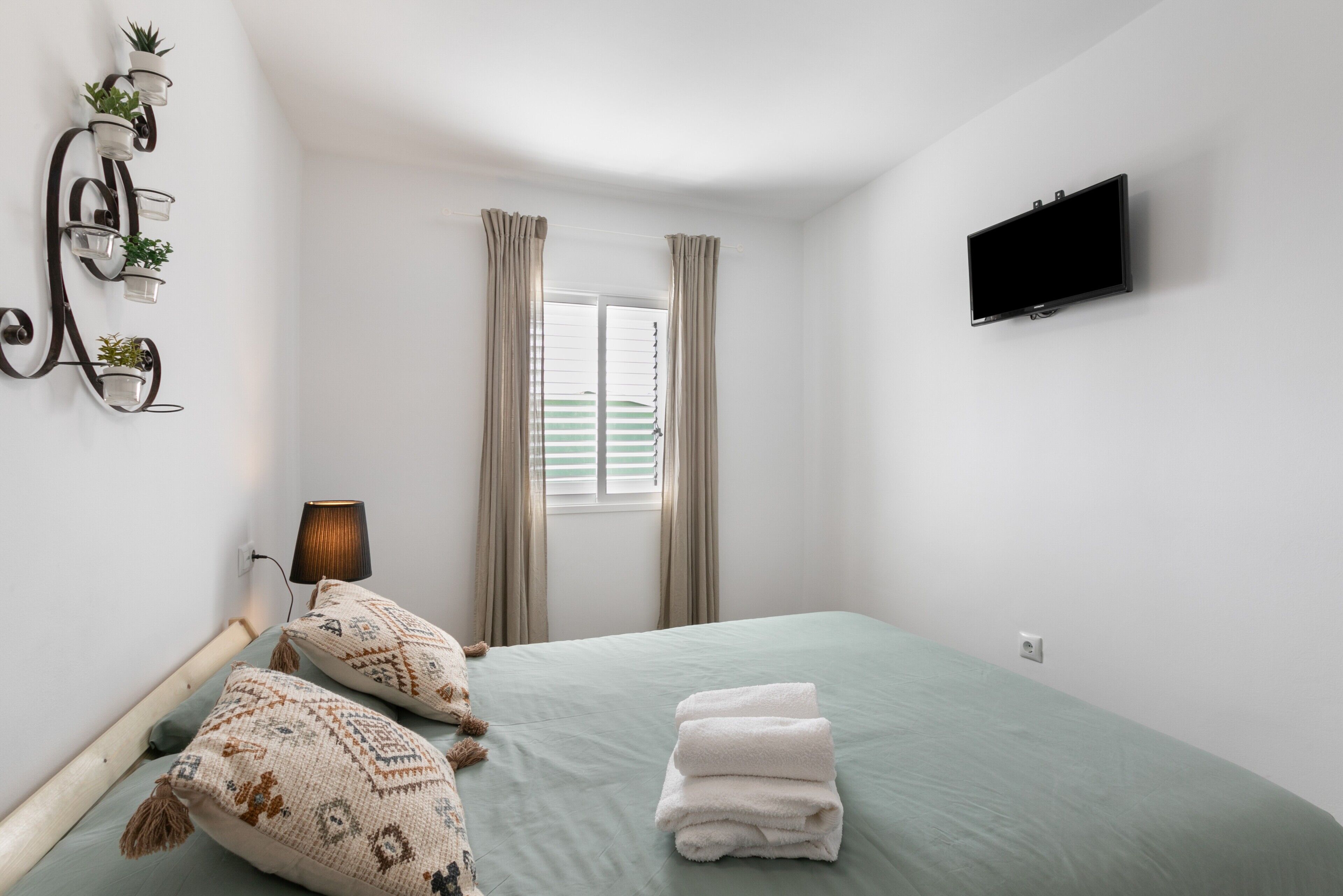 Une chambre à coucher avec un lit, des oreillers, une télévision, une fenêtre avec des rideaux et une étagère à plantes fixée au mur.