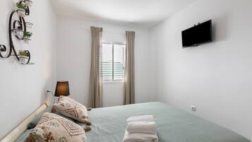 Une chambre à coucher avec un lit, des oreillers, une télévision, une fenêtre avec des rideaux et une étagère à plantes fixée au mur.