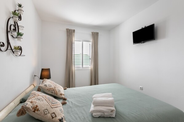 A bedroom with a bed, pillows, a TV, a window with curtains, and a wall-mounted plant shelf.