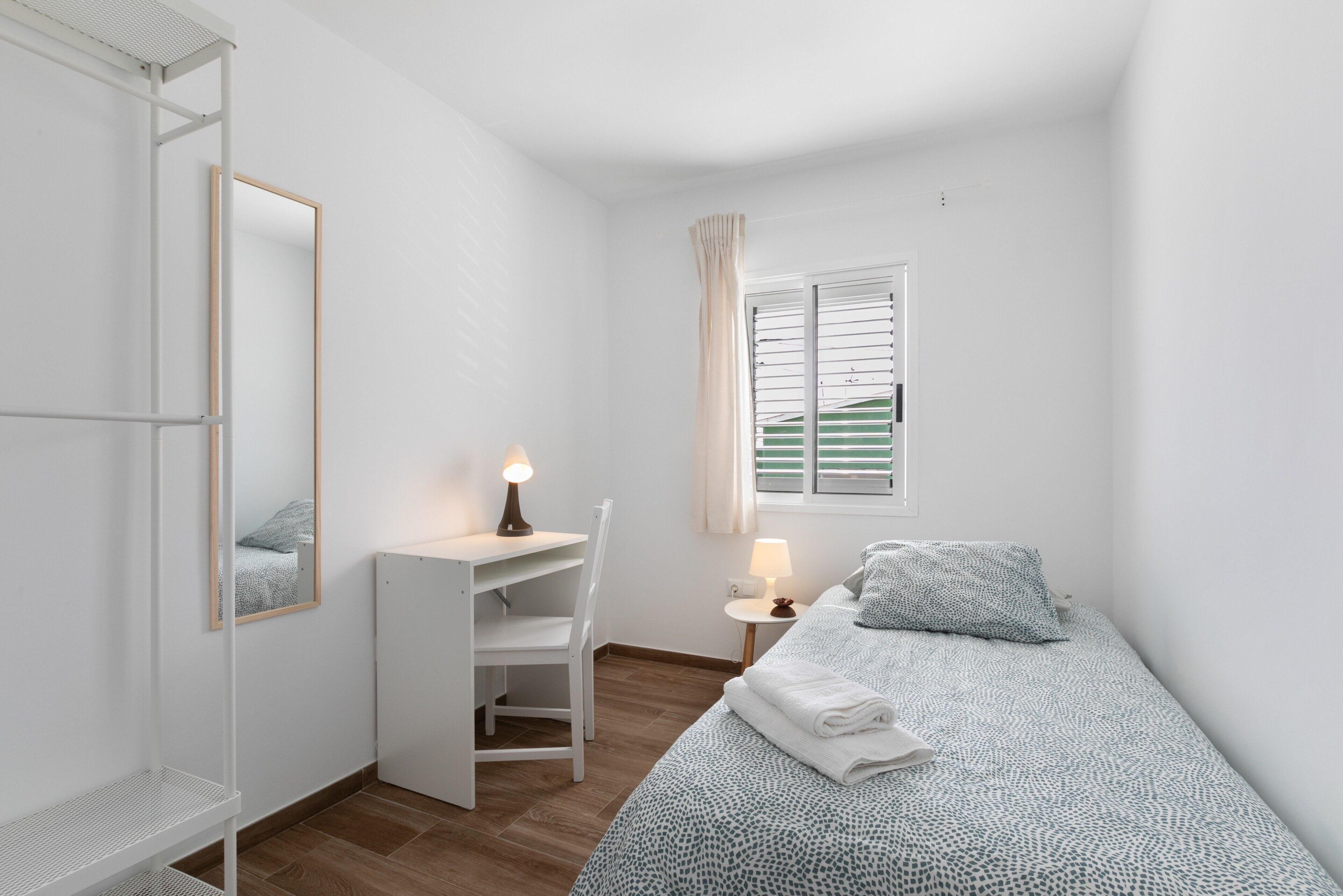 A bedroom with a bed, a desk, a chair, a mirror, a window with curtains, and a radiator.