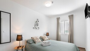 Ein ordentlich eingerichtetes Schlafzimmer mit einem Bett, Nachttischlampen, einem am Wand montierten Fernseher und einem Fenster mit Vorhängen.