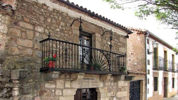 Edifici de pedra amb una porta de fusta, un balcó amb barana de ferro forjat i una petita planta en test.