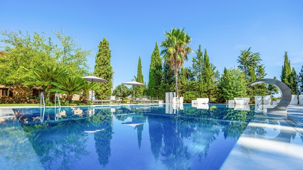 Une piscine aux eaux cristallines, bordée de chaises longues et d’ombrelles, entourée d’arbres et surplombée d’un ciel dégagé.