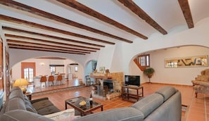 Amplia sala de estar con vigas de madera a la vista, chimenea, televisor y comedor al fondo.