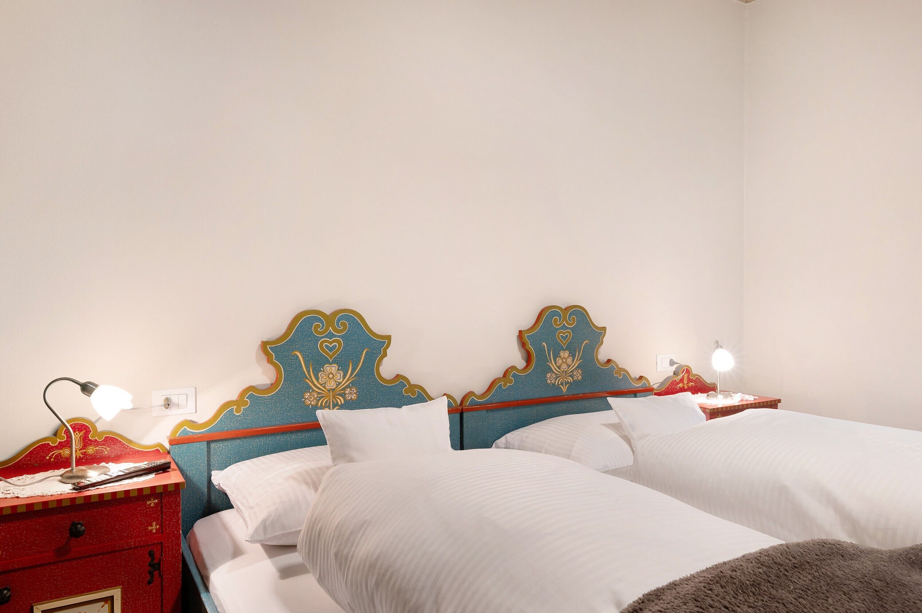 Ein Doppelbett mit weißen Leinen, ein roter Nachttisch mit Lampe sowie ein blaues Kopfteil mit Verzierungen.