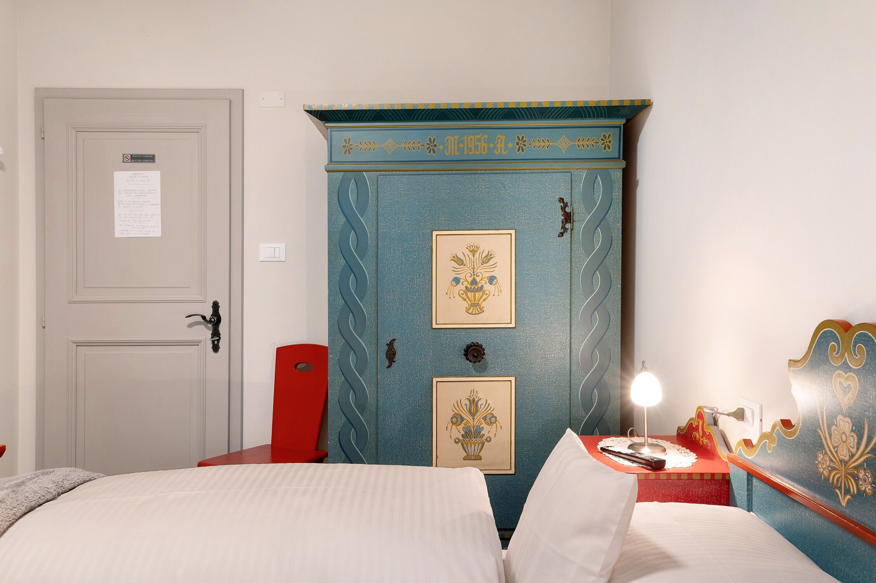 A hotel room with a bed, a nightstand with a lamp, a dresser with decorative panels, and a door with a sign.