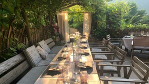 Una larga mesa de comedor dispuesta para una comida, rodeada de bancos y sillas de madera, con vistas a un jardín.