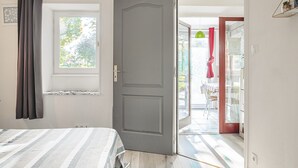A room with a bed, a window with curtains, a door leading to an outdoor area with a table and chairs, and a shelf with items on the wall.