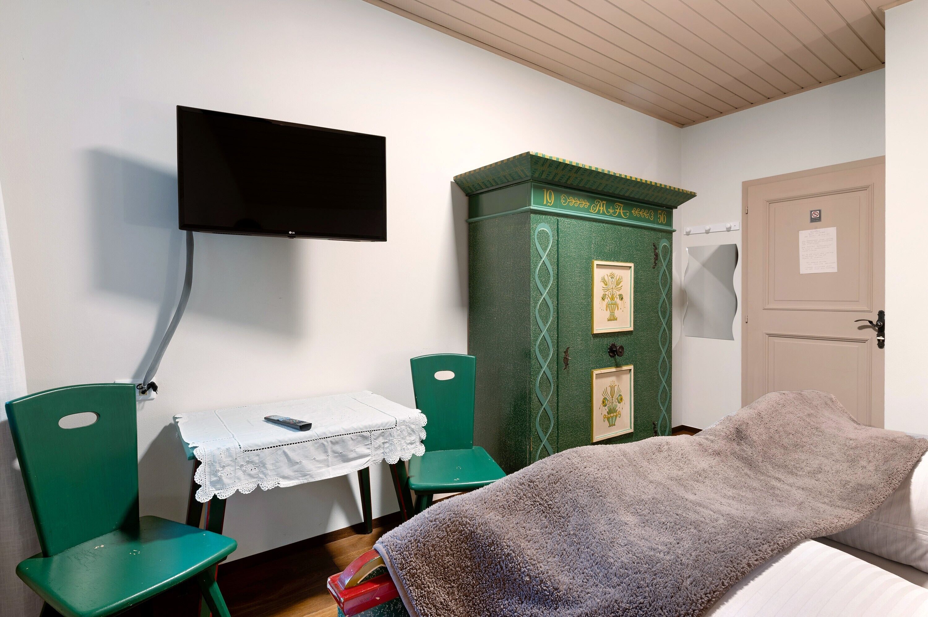 A room with a bed, a desk with a chair, a television mounted on the wall, and a green cabinet with a decorative top.