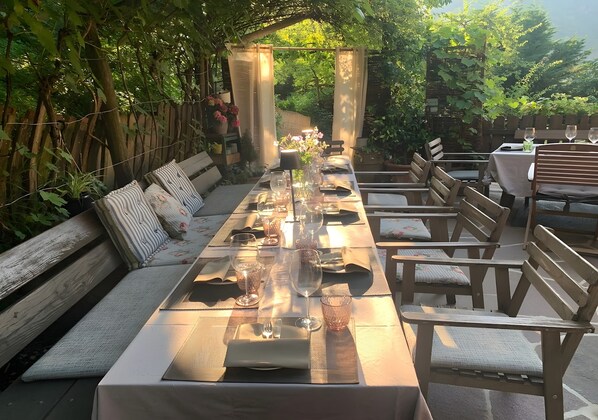 Une longue table à manger, dressée pour un repas, entourée de bancs et de chaises en bois, avec vue sur le jardin.
