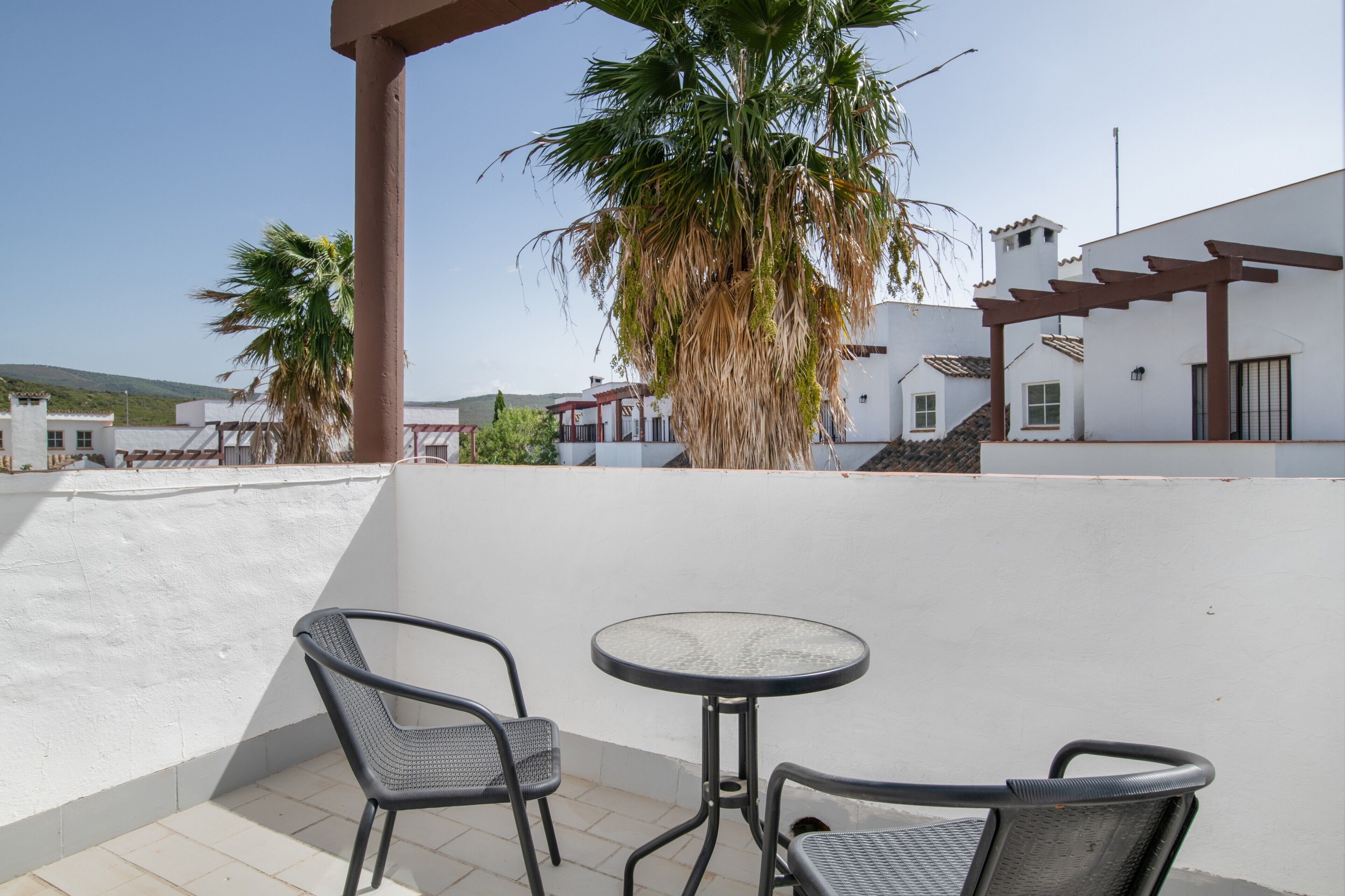 A terrace with two chairs and a table, a palm tree, and a white building with a red roof.