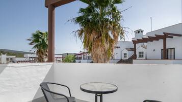 A terrace with two chairs and a table, a palm tree, and a white building with a red roof.