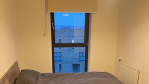 A bedroom with a large bed, a window with a view of buildings, a radiator, and a ceiling light.