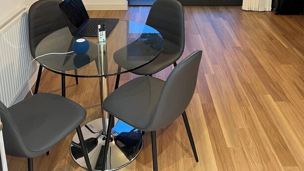 A modern room with a glass table, four chairs, a laptop, and a view of a cityscape through large sliding doors.