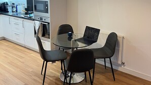 A modern kitchen with white cabinets, a microwave, and a kettle. There is a dining area with a glass table and black chairs.