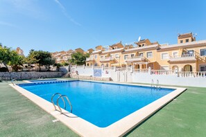 Una piscina rectangular con dos tableros de salto, rodeada por un césped verde y una hilera de edificios color beige con balcones.