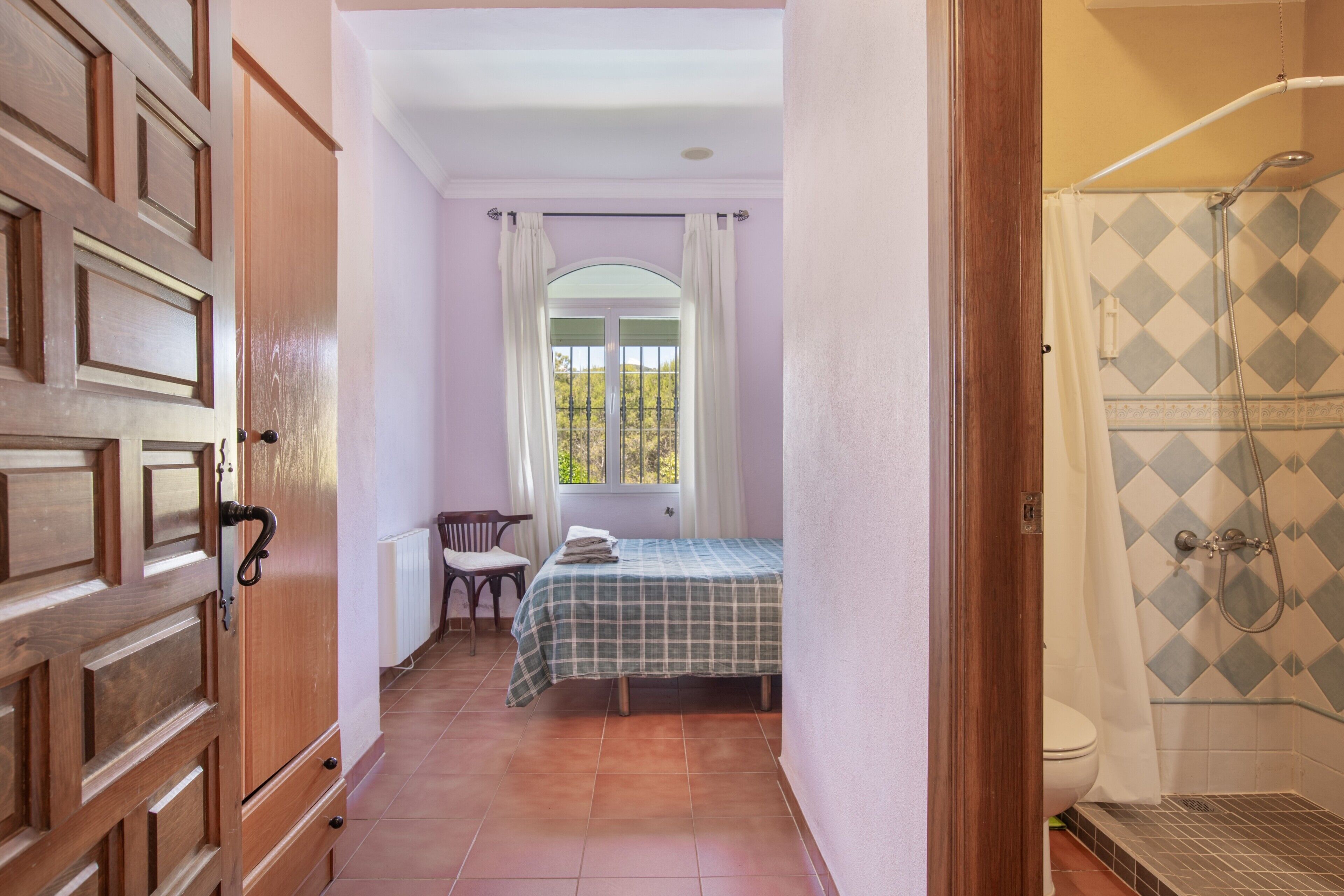 A bedroom with a bed, a window with curtains, a wooden door, and a shower area.