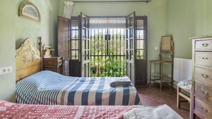 Une chambre à coucher comprenant un lit, une commode, une table de chevet, une fenêtre donnant sur un espace verdoyant et une porte menant à un balcon.