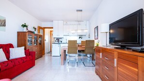 Une cuisine moderne avec des meubles blancs, un coin repas avec des chaises en osier, un canapé rouge et un téléviseur à écran plat.