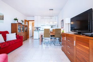 A modern kitchen with white cabinets, a dining area with wicker chairs, a red sofa, and a flat-screen TV.