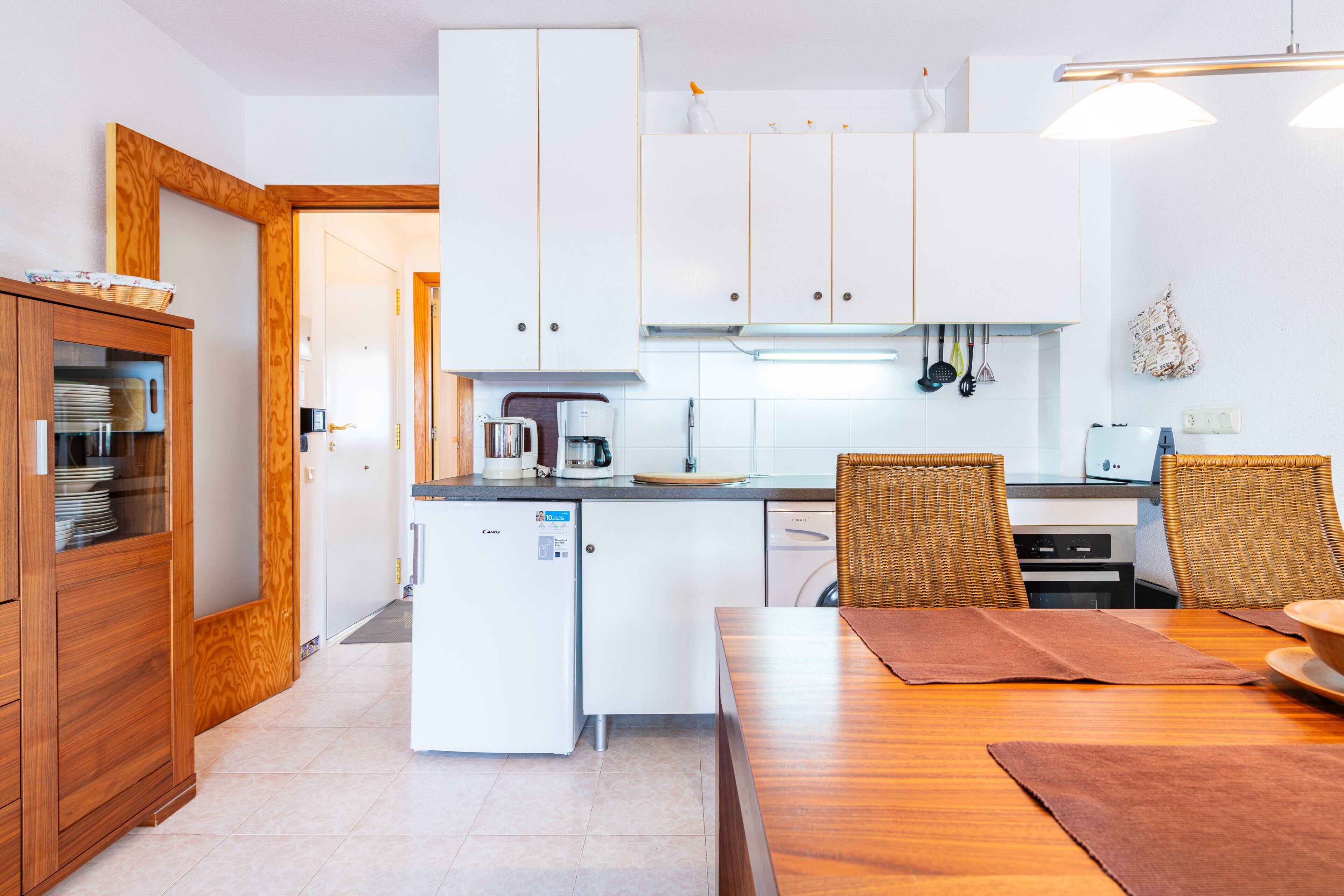 A compact kitchen with white cabinets, a small refrigerator, a coffee maker, and a dining area with a wooden table and wicker chairs.