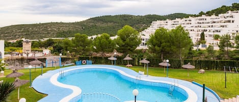 A swimming pool with a wavy edge, surrounded by a fence, with a grassy area and trees in the background.