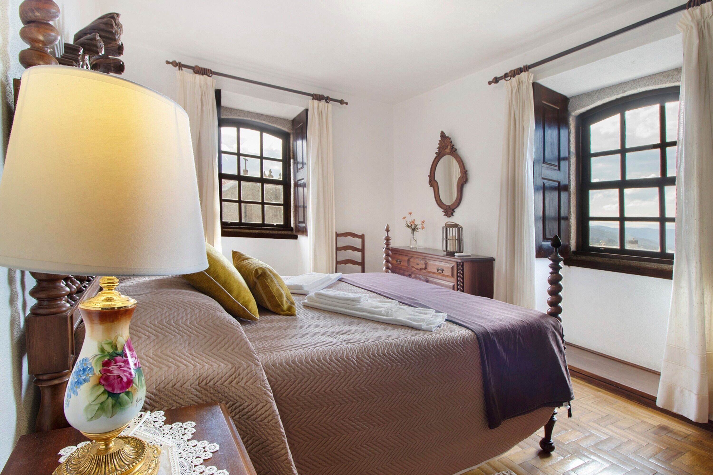 Een slaapkamer met een bed, nachtkastje, lamp, dressoir en raam met gordijnen.