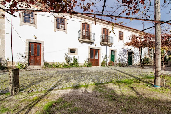 A white building with multiple doors and windows, a cobblestone courtyard, and a tree with bare branches.