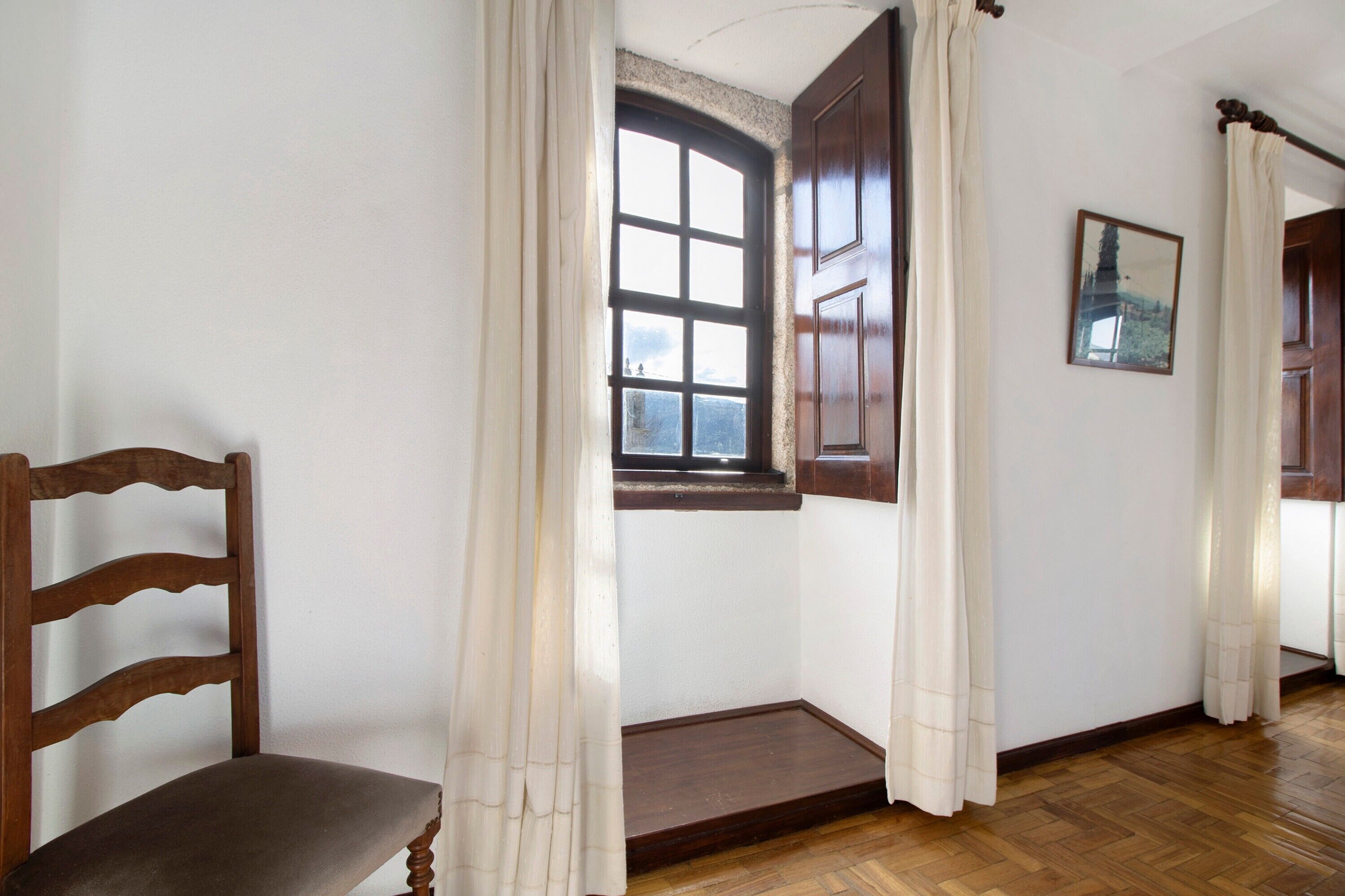 A room with a wooden chair, a window with curtains, a wooden door, and a framed picture on the wall.