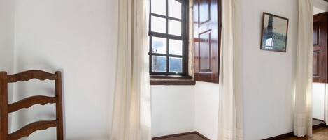 A room with a wooden chair, a window with curtains, a wooden door, and a framed picture on the wall.
