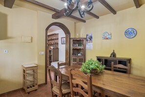 A dining area with a wooden table, chairs, a potted plant, a wall-mounted calendar, and a shelf with glassware.
