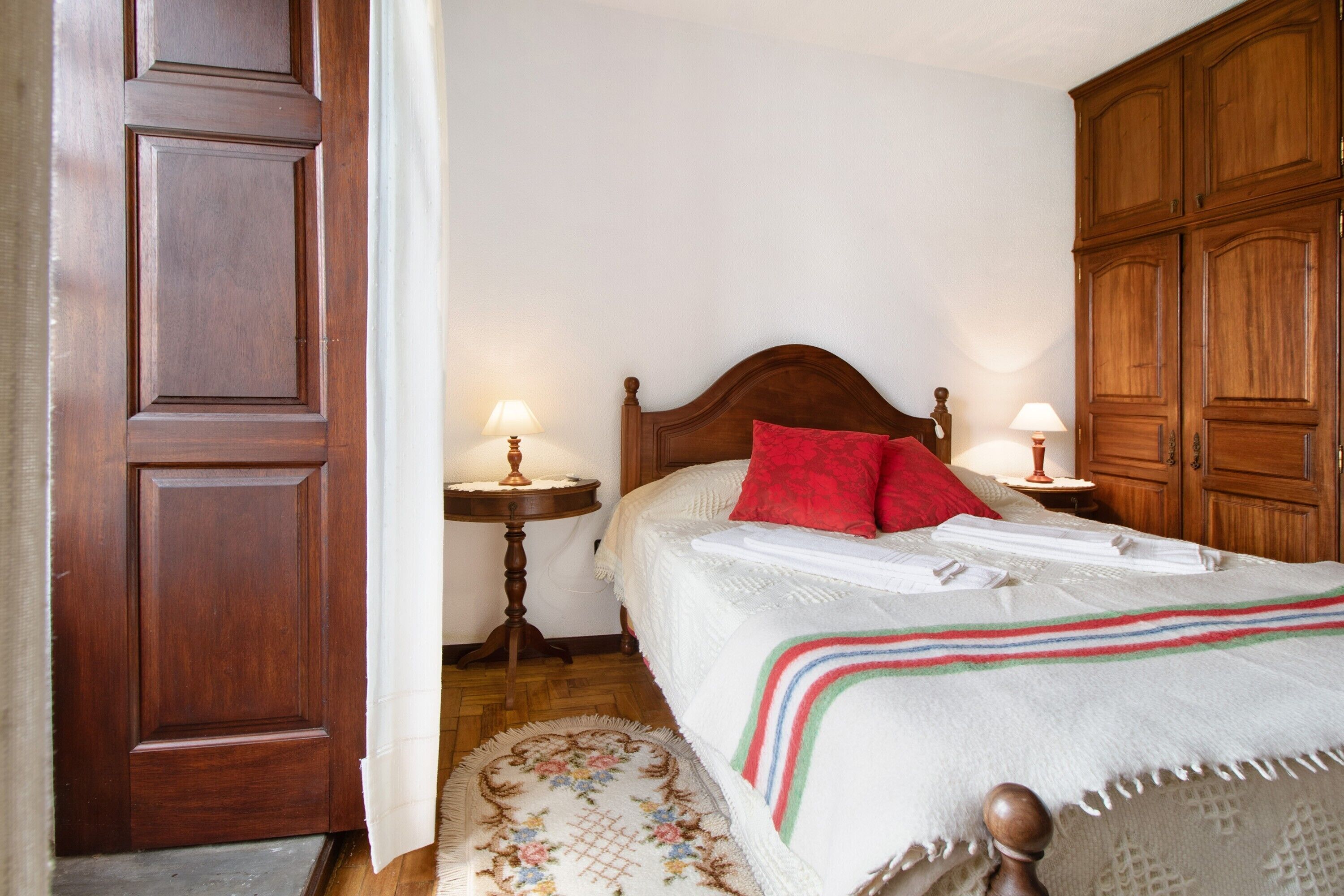 Un dormitorio con una cama de madera, dos mesitas de noche con lámparas, un armario de madera y una alfombra estampada.