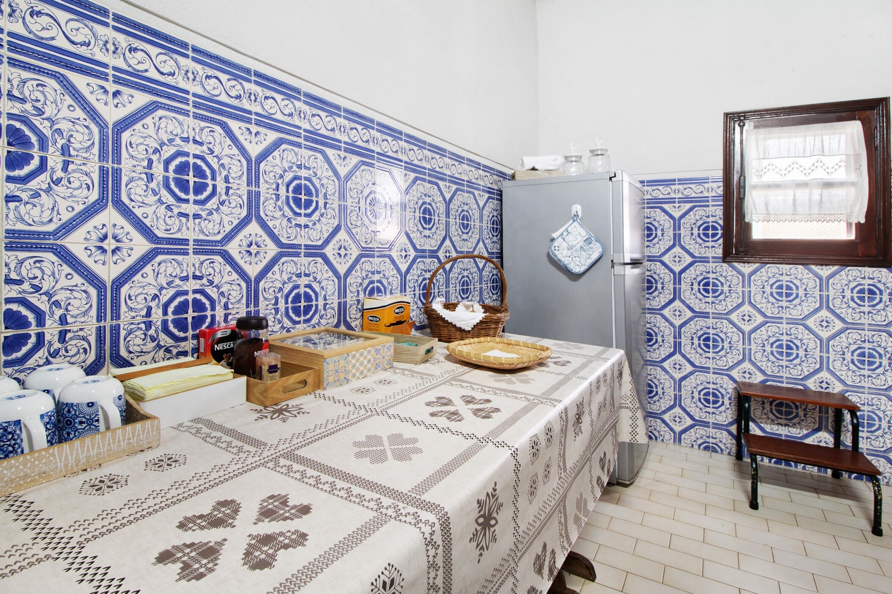 Una cucina con piastrelle a motivi blu e bianchi, un frigorifero, un tavolo di legno con tovaglia e una finestra con tenda.